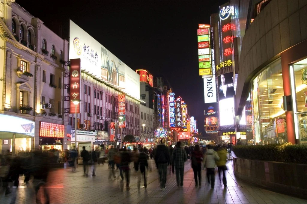 Crowded nighttime shopping street with neon signs and storefronts, showing an urban retail discovery environment.