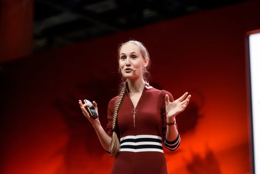 Ashley Dudarenok presenting on stage with a headset microphone against a red background